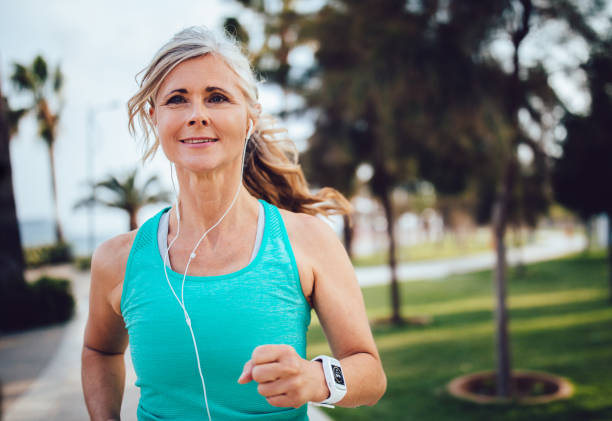 Active mature woman with headphones and smartwatch running in park Athletic senior woman with smartwatch jogging and listening to a podcast in the park healthy lifestyle women outdoors athlete stock pictures, royalty-free photos & images
