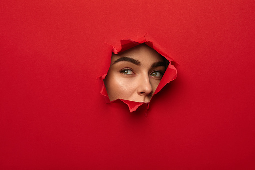 Young female face looking out of hole torn in red paper.