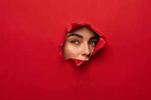 Young female face looking out of hole torn in red paper.