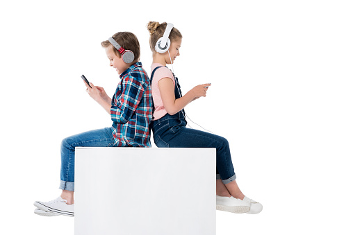 children using smartphones in headphones while sitting on cube together isolated on white