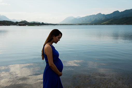 Side view of a young latin pregnant woman holding belly and looking down in a horizontal medium shot outdoors next to lake.