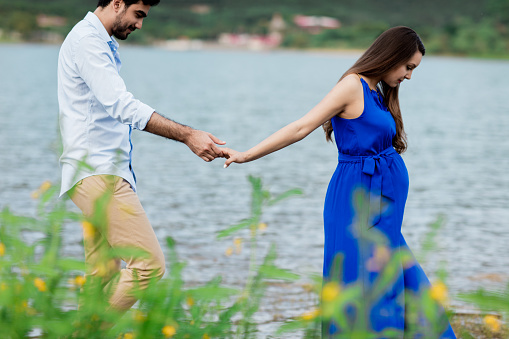 Side view of young expecting parents walking next to a lake and holding hands in a horizontal medium shot outdoors.