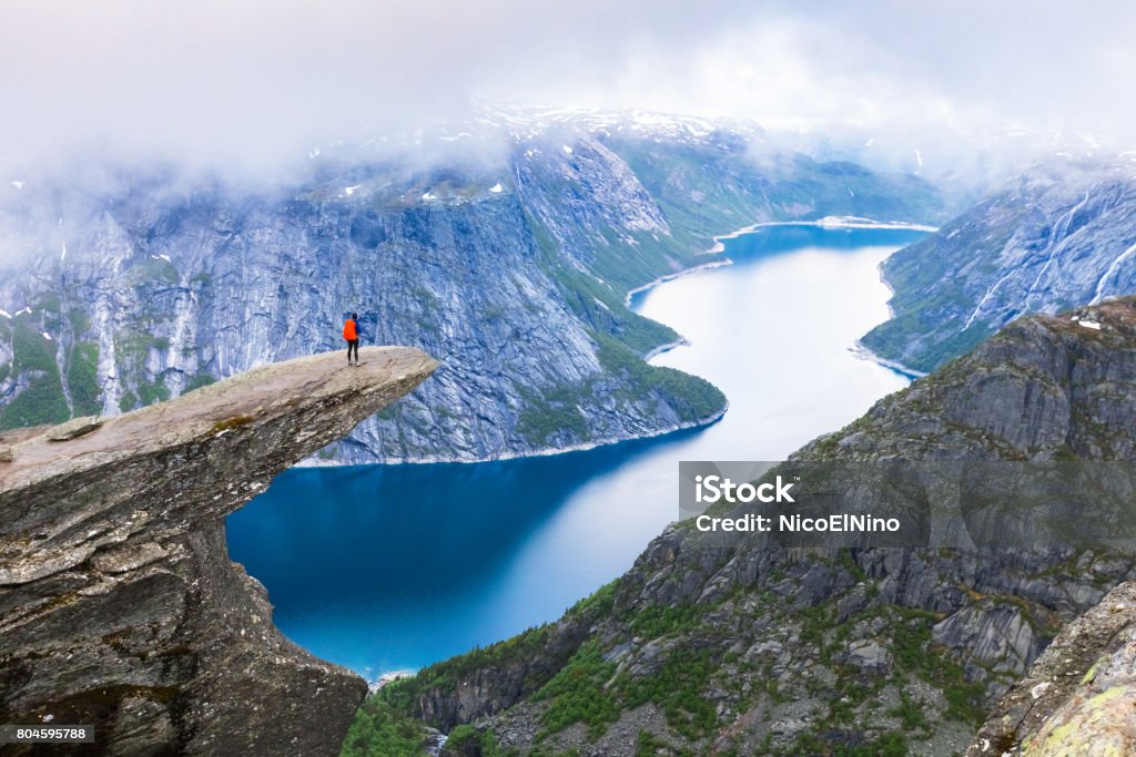 Hiker standing on Trolltunga with panoramic view of lake, Norway Female hiker standing on Trolltunga (troll tongue) famous rock high above a lake with panoramic view of the valley, popular tourist adventure trek in Norway Norway Stock Photo