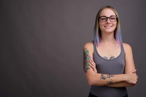 Studio shot of young beautiful woman with multi colored hair and tattoos wearing gray tank top against gray background horizontal shot