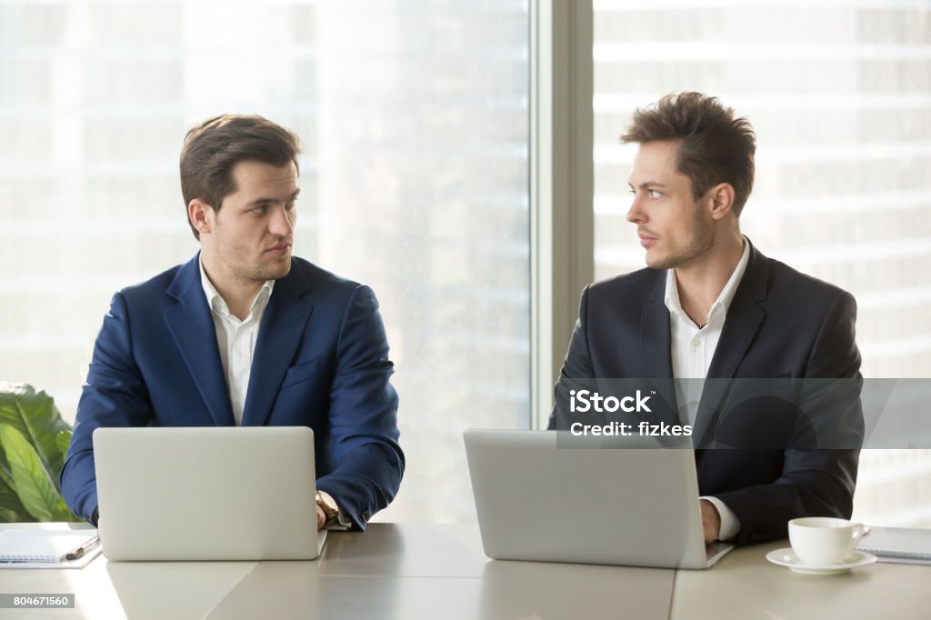 Two confident businessmen dislike each other, business competition and rivalry Two confident businessmen sitting at office desk with laptops, looking at each other with hate dislike, rivals accepting challenge, business competition, team rivalry at work, competitors behavior Meeting Stock Photo