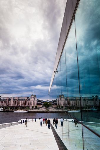 Oslo, Norway - 13 June, 2015: Wide angle image depicting the modern exterior of the opera house in Oslo, Norway. Crowds of tourists explore its white marbled walkways. The Oslo fjord can be seen in the foreground. Vertical colour image with copy space.