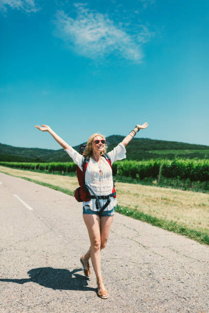 Woman on the road trip. Walking alone down the road with backpack on her back and with raised arms .