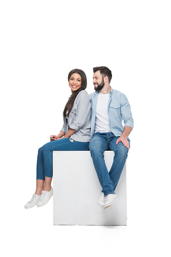 multiethnic happy couple sitting on cube together isolated on white