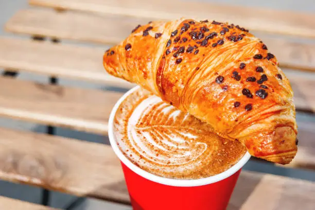 Red paper cup with coffee and chocolate croissant. Coffee to go. Tasty hot beverage on wooden table in sunny day. Outdoors meal.