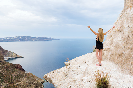 Horizontal color rear view image of beautiful woman with arms raised standing on cliff above the Mediterranean sea. Freedom, holidays & harmony.