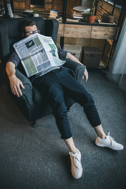 istock Tired young man in eyeglasses sleeping with newspaper at home 805124026