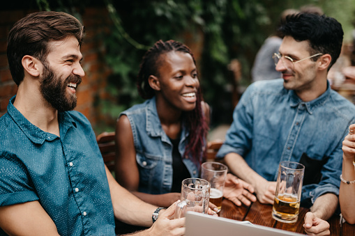 Multi-ethnic group of people sitting at the pub, drinking beer, using laptop, having fun. Colleagues gathered at the bar after work to relax. Five friends at the fastfood restaurant enjoying their free time together.