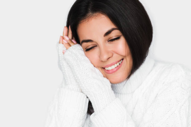 Portrait of beautiful young happy dreamy woman with closed eyes in white sweater on light grey background. Pretty brunette model with beautiful face, studio portrait. Healthy mood. stock photo