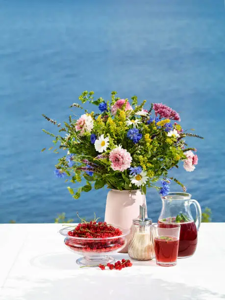 Bouquet of wild flowers on table with red currants and limonade. View behind over the sea