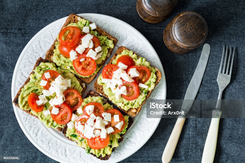 Avocado, tomato and cheese on toasted bread. Healthy snack, appetizer Avocado toast. Healthy toast with avocado mash, cherry tomatoes and crumbled feta cheese on a plate. Table top view Avocado Stock Photo