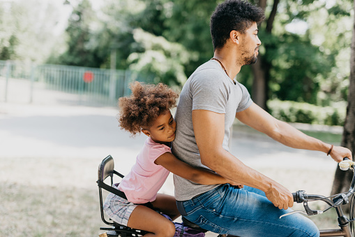 Carefree African American father and daughter cycling in the park. Mixed race family riding a bike together through the urban part of the city. A loving parent cycles around the block with his kid sitting in the children's seat.