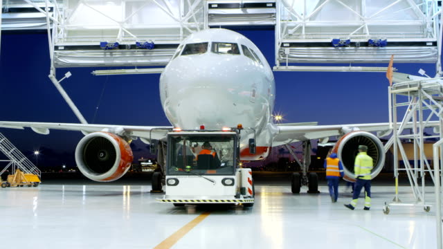 Time-Lapse of a Aircraft Maintenance Hangar Where New Airplane is Toed by a Pushback Tractor/ Tug onto Landing Strip. Crew of Mechanics, Engineers and Drivers Works Busily.