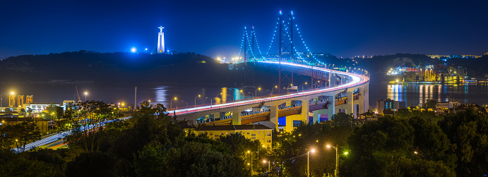 The iconic span of the 25 de Abril Bridge illuminated by the lights of the suspension cables and the zooming traffic crossing from Almada to Lisbon, Portugal’s vibrant capital city.