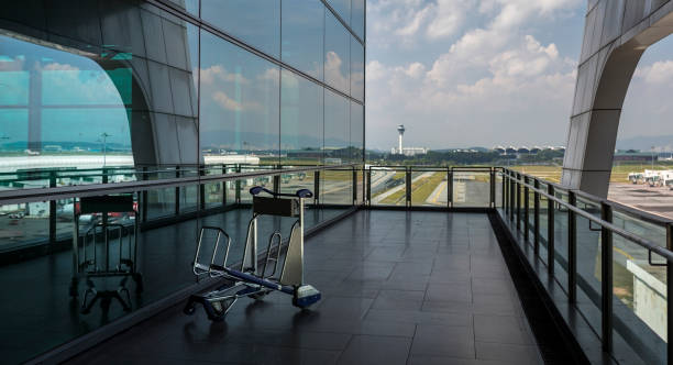 Luggage cart at a modern airport in Asia In a modern outdoor corridor of an international airport, an empty luggage cart stands by itself with glass window reflection behind and an airport traffic control tower in the background. klia airport stock pictures, royalty-free photos & images