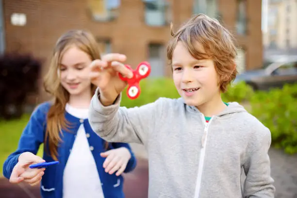 Photo of Two funny friends playing with fidget spinners on the playground. Popular stress-relieving toy for school kids and adults.