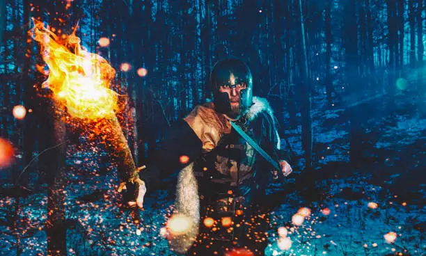 Male hunter is armed with sword and a flaming torch as he walks through a forest at night.