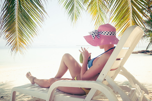 Young and sensual hipster girl is sitting on white plastic easy chair under the palm tree. She is holding coconut with straw in her hands and drinking from it. All around her is amazing beach, white sand, turquoise water and clear sky. She is wearing yellow bottom of bikini, blue top of bikini and pink hat with white and black ribbon. Next to the easy chair on sand is big red bag with white stripes. Sunny warm day is great to get some tan and relax next to the water or under palm tree.  Peaceful environment and heavenly island is amazing combination for enjoyment and relaxing.