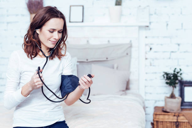 Involved young woman using tonometer at home Testing new health care device . Pleasant skilled young woman sitting on the bed at home and measuring blood pressure while holding tonometer picture of protractor stock pictures, royalty-free photos & images