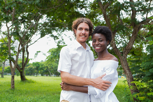 Wedding portrait of happy young mix-raced couple, African-American woman and Caucasian man, embracing, looking at camera and smiling in summer park