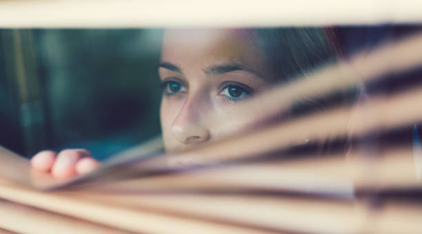 Unhappy woman looking through the window Scared woman at home looking through the shutters sheepish look stock pictures, royalty-free photos & images