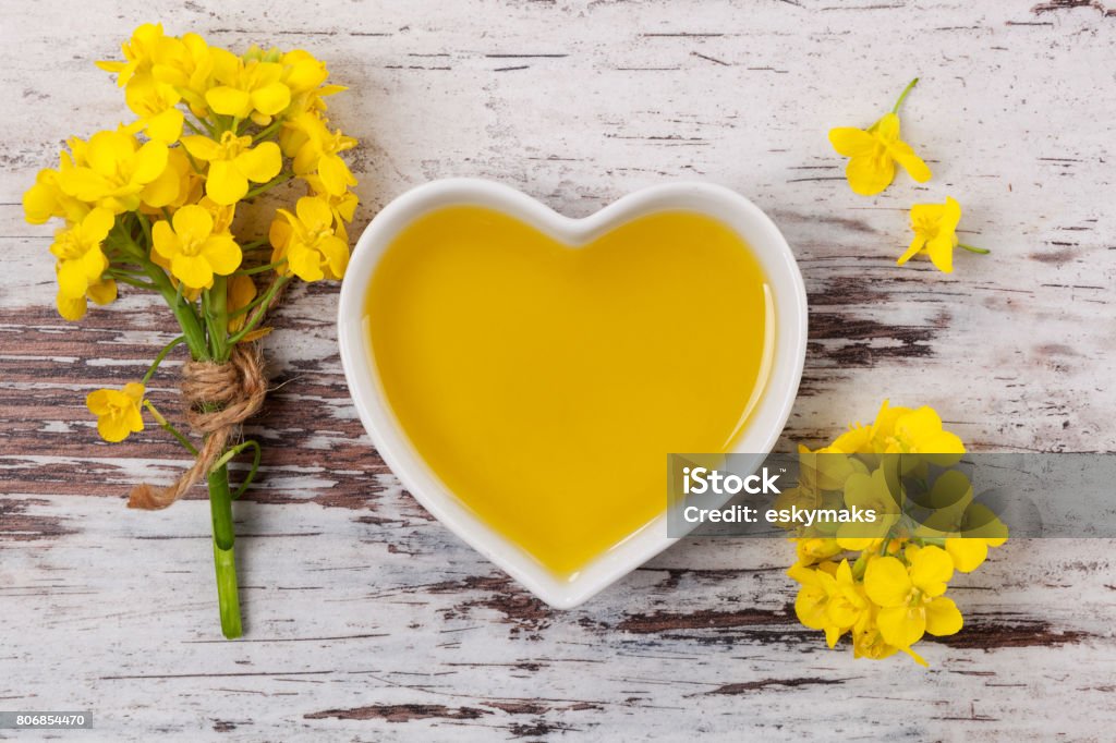 Rapeseed oil and flower. Rapeseed oil in heart shaped bowl and flowers on wooden background, top view. Canola Stock Photo