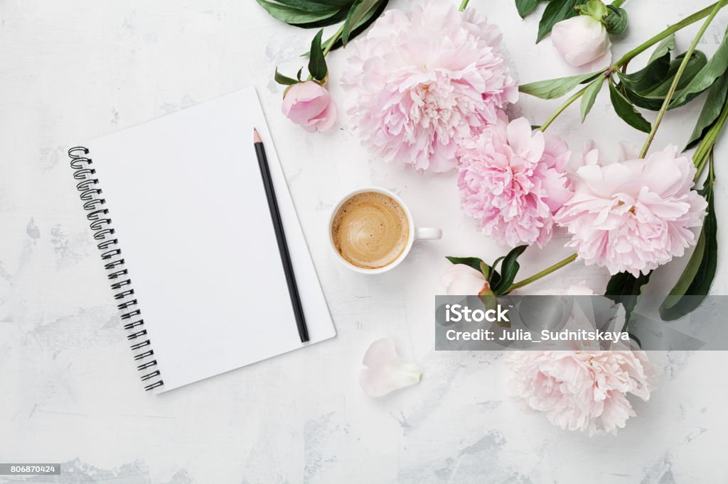 Morning coffee mug for breakfast, empty notebook, pencil and pink peony flowers. Flat lay. Woman working desk. Morning coffee mug for breakfast, empty notebook, pencil and pink peony flowers on white stone table top view in flat lay style. Woman working desk. Desk Stock Photo