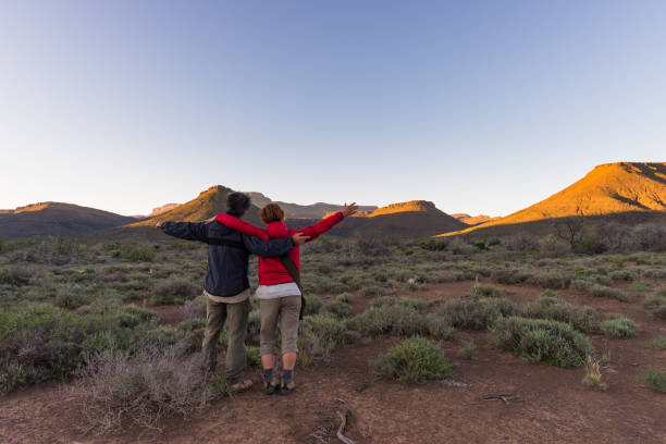 hugging couple with outstretched arms watching the stunning view of the karoo national park at sunset, travel destination in south africa. traveling people and adventure. - landscape panoramic kalahari desert namibia imagens e fotografias de stock