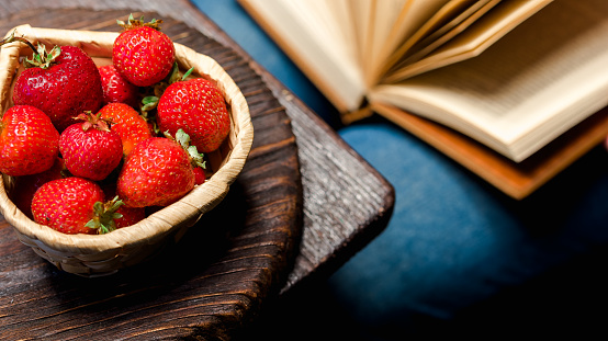 Fresh strawberries in basket  on the table corner. Person sitting at the table holding open book on her knees. Reading and eating concept