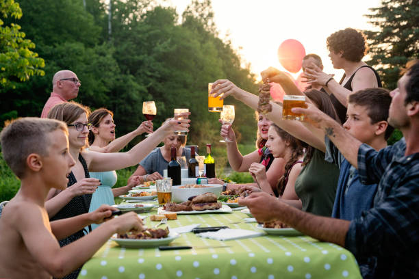 Big family barbecue gathering at sunset, summer outdoors. Celebration time for this three generation real family outdoors in summer. Big dinner is ready, cheers everyone! Horizontal waist up shot with copy space. This was taken in Quebec, Canada. big family table stock pictures, royalty-free photos & images