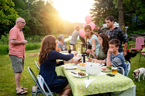 Big family barbecue gathering at sunset, summer outdoors. Celebration time for this three generation real family outdoors in summer. Big dinner is ready, people coming to the table. Horizontal full length shot with copy space. This was taken in Quebec, Canada. big family sunset stock pictures, royalty-free photos & images