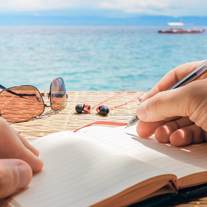 caucasian man is writing some idea, message or letter in his notepad by pen in his right hand while he sitting on the beach of tropical sea with boat at sunny day