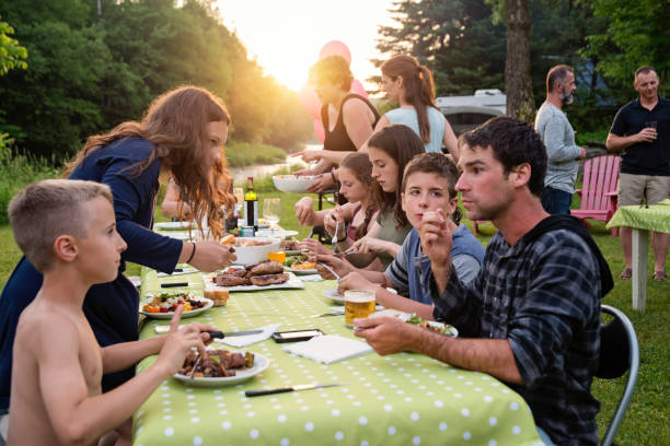 Big family barbecue gathering at sunset, summer outdoors. Celebration time for this three generation real family outdoors in summer. Big dinner is ready, people coming to the table. Horizontal waist up shot with copy space. This was taken in Quebec, Canada. big family sunset stock pictures, royalty-free photos & images