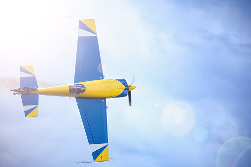 A small sport plane flying in the sky. A sun flare in the corner. Color of the plane is blue and yellow. a pilot can be seen through cabin's bulb.