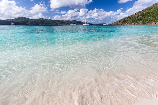 Waves gently wash up on a beautiful white sand beach of Guana island in the British Virgin Islands in the Caribbean. Several anchored sailboats are in the background.