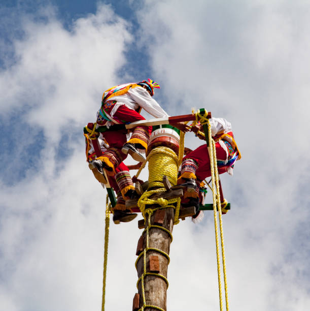 A typical mexican indian show where four men wearing typical colorful clothes jump from a pole tied to a yellow rope under the rythm of a musician.