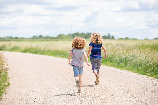 Rear view of two young girls (sisters) running down a rural gravel driveway on a summer day. The older sister is in the lead and has curly blonde hair. The younger sister is nearest the camera and has curly red hair.