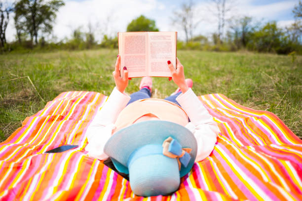 Young woman with blue hat dressed casually reads orange old book lying down on a colourful blanket in the park. Traditional reading.