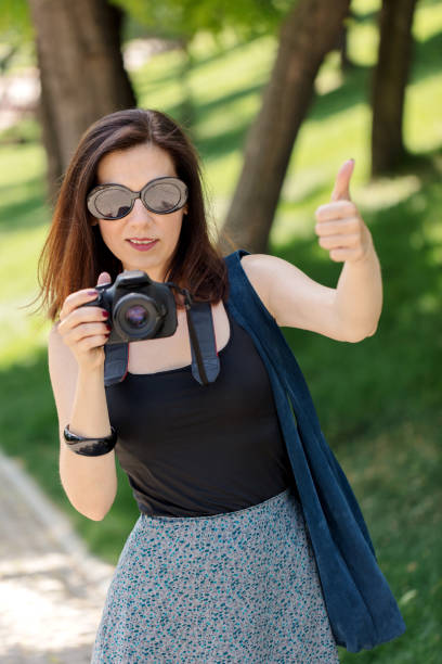 Young woman (photographer, tourist) holds a camera and shows a thumb gesture (pollex, digitus primus). Young woman (photographer, tourist) holds a camera and shows a thumb gesture (pollex, digitus primus). Selective focus. pollex stock pictures, royalty-free photos & images