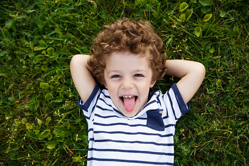 Curly-haired boy in striped t-shirt with hands behind head lying on grass smiling and sticking out tongue.