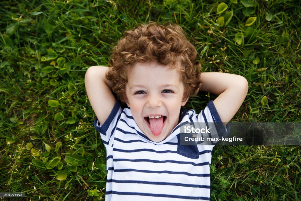 Curly-haired boy lying on grass Curly-haired boy in striped t-shirt with hands behind head lying on grass smiling and sticking out tongue. Child Stock Photo