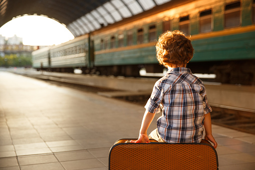 Young boy wearing check shirt sitting on brown suitcase at railroad station exposed to light.