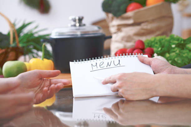 istock Close-up of human hands pointing into note book copy space area in the kitchen. Friends having fun while choosing menu or making online shopping. So much ideas for tasty cooking. Vegetarian, healthy meal and friendship concept 807332480