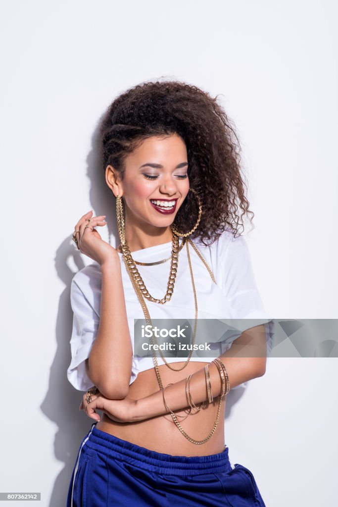 Vertical portrait of happy young latin woman in chola style Studio portrait of chola style young latin woman wearing gold jewellery and tracksuit pants, laughing against white background. 20-24 Years Stock Photo
