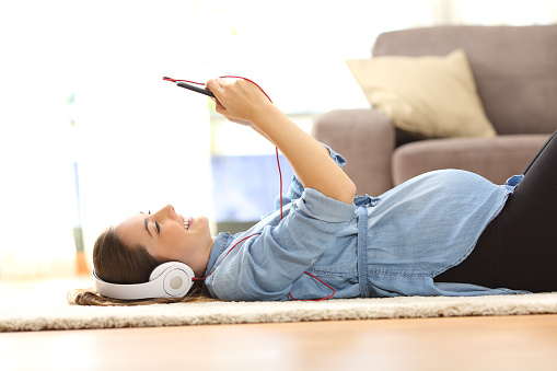 Side view of a pregnant woman listening music with headphones and a smart phone lying on the floor at home