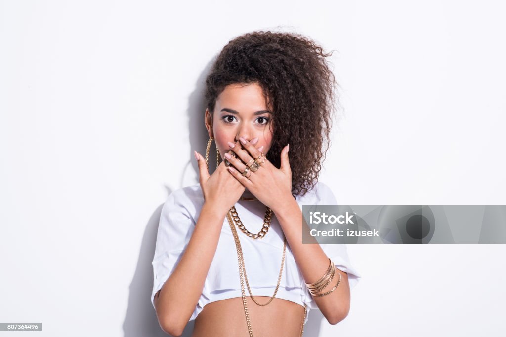 Surprised chola style young woman Studio portrait of surprised chola style young latin woman wearing gold jewellery, staring at camera with hands on her mouth. White background. Jewelry Stock Photo